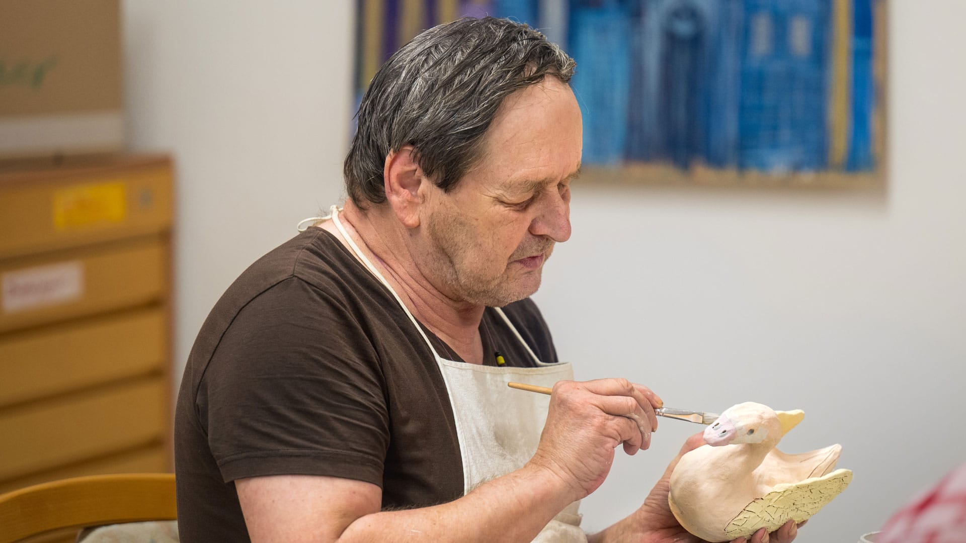 Ein älterer Mann malt konzentriert eine weiße Figurenstatue. Er trägt ein braunes T-Shirt und eine Schürze.