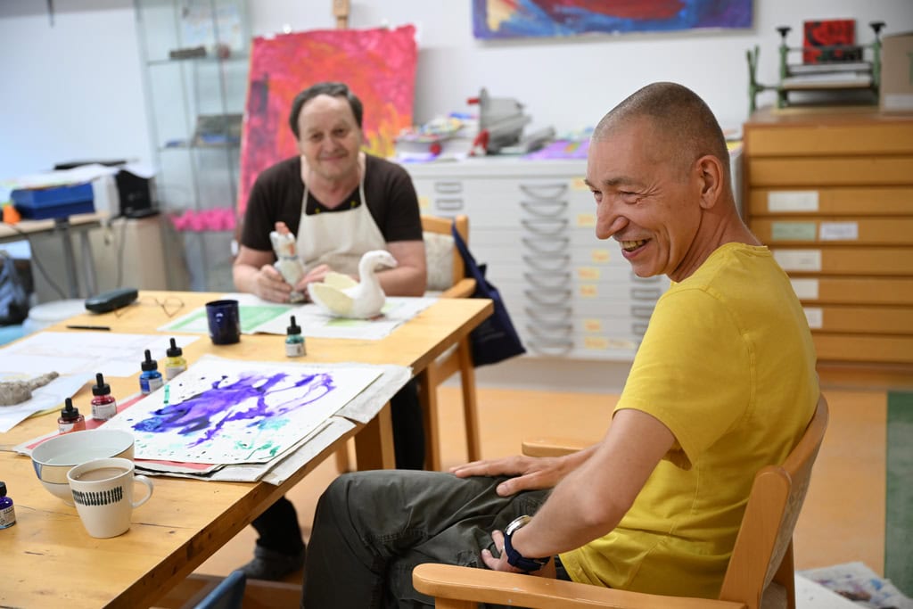 Zwei Männer lachen in einem Kunstraum. Einer malt, der andere trägt ein gelbes T-Shirt und sitzt lachend.